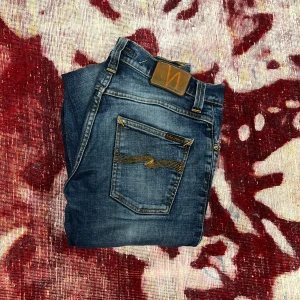 Blå jeans från Nudie Jeans - Säljer ett par snygga blå jeans från Nudie Jeans med klassisk femficksdesign och orangea sömmar. Jeansen har en straight passform och är tillverkade i slitstarkt denim. Perfekta för en avslappnad stil. Dom har ett litet hål i höger framfickan som ni ser på sista bilden