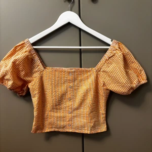 Orange- och vitrutad topp från Edited - Säljer en söt orange- och vitrutad topp från Edikted. Toppen har puffiga korta ärmar och knappar framtill. Perfekt för en somrig look! 🌼