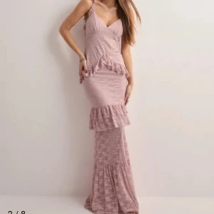 Rosa spetsklänning med volanger - Elegant rosa långklänning i spets med volangdetaljer och smala axelband. Klänningen har en v-ringad design och en öppen rygg med knytning. Perfekt till bal! (Prislappen är kvar) 