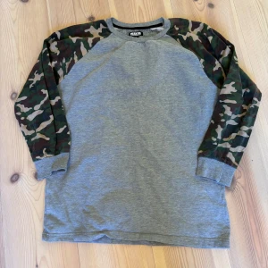 Grå tröja med camouflageärmar från H&M - Säljer en grå tröja från H&M med coola camouflageärmar. Perfekt för en avslappnad stil. Tröjan har långa ärmar och en bekväm passform. Passar bra till jeans eller joggers.