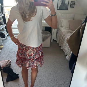 Mönstrad kortkjol i rött och blått - Säljer en färgglad kortkjol med ett livligt mönster i rött, blått och vitt. Kjolen har volanger som ger en lekfull touch. Perfekt för sommarens äventyr!