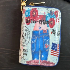 Färgglad plånbok med tryck - Säljer en unik plånbok med ett färgglatt tryck av blommor, jeans och amerikansk flagga. Plånboken har en dragkedja och en praktisk handledsrem. Perfekt för att hålla ordning på kort och kontanter. Plånboken har aldrig använts 