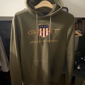 Olivgrön hoodie från GANT - Säljer en snygg olivgrön hoodie från GANT med broderad logga och text på framsidan. Hoodien har en klassisk känguruficka och justerbar huva med dragsko. Perfekt för en avslappnad stil.
