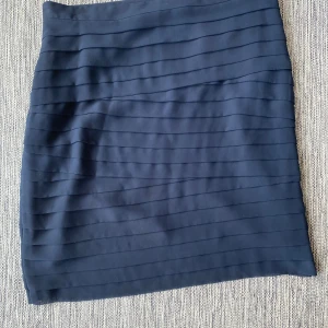 Mörkblå kjol från GANT - Snygg mörkblå kjol från GANT med ett stilrent randigt mönster. Kjolen är tillverkad i polyester och elastan för en bekväm passform. Perfekt för en elegant look.  Marinblå volangkjol i strl 46 från Gant.  Kjolen har en underkjol.  Sparsamt använd och i fint begagnat skick förutom att det finns några lösa trådar på ena sidan.  Mått Midjan: ca 48 cm Total längd: ca 62 cm  Polyester och elastan  Kommer från ett djur och rökfritt hem.