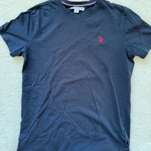 Mörkblå t-shirt från U.S. Polo Assn. - Säljer en stilren mörkblå t-shirt från U.S. Polo Assn. med ett rött broderat emblem på bröstet. T-shirten har en klassisk passform och är tillverkad i mjukt bomullsmaterial. Perfekt för en avslappnad look. Storlek M