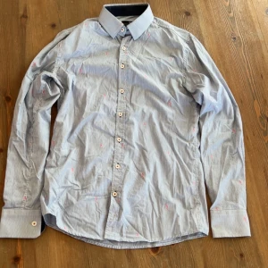 Blå skjorta från Savvy Citizen - Snygg blå skjorta från Savvy Citizen med diskret randigt mönster och små broderade detaljer i rosa. Skjortan har långa ärmar och knappar framtill. Perfekt för en stilren look.