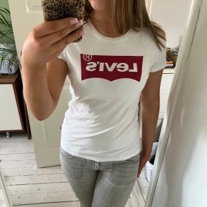 Vit t-shirt från Levi's - Säljer en klassisk vit t-shirt från Levi's med det ikoniska röda logotyptrycket på bröstet. T-shirten har en rund hals och korta ärmar, perfekt för en avslappnad stil. Köpt i USA, använt fåtal gånger.