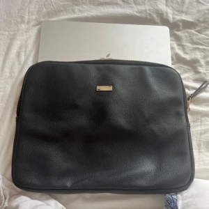 Datafodral - Snygg och stilren svart datorväska i skinn med dragkedja. Perfekt för att skydda din laptop med stil. Väskan har en enkel design med en liten metallplatta som detalj.