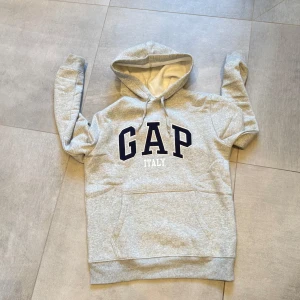 Grå hoodie från GAP - Säljer en klassisk grå hoodie från GAP med texten 'GAP ITALY' i svart och vitt på bröstet. Tröjan har en stor ficka framtill och justerbar huva med snören. Perfekt för en avslappnad stil. Bara att höra av sig vid funderingar!🤩