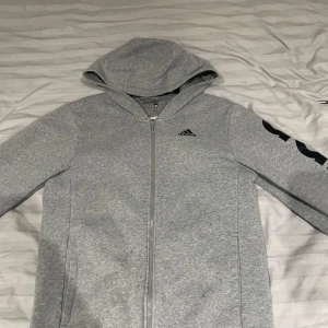 Grå hoodie från Adidas - Säljer en stilren grå hoodie från Adidas med dragkedja och huva. Tröjan har långa ärmar och Adidas-loggan tryckt i svart på ärmen och bröstet. Perfekt för en avslappnad stil.