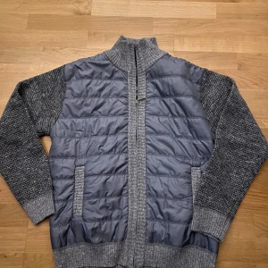 Grå quiltad cardigan jacka - Snygg grå cardigan jacka med quiltade detaljer och dragkedja framtill. Ärmarna och baksidan är stickade vilket ger en skön och varm känsla. Perfekt för kyligare dagar.