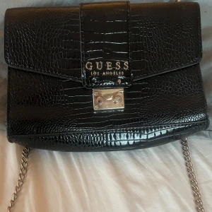 Svart axelväska från Guess - Snygg svart axelväska från Guess med krokodilpräglat mönster och silverdetaljer. Väskan har en kedjerem och ett praktiskt innerfack med dragkedja. Perfekt för att ge din outfit en lyxig touch. Har tappat en nit vid framsidan men inget som påverkar användningen.