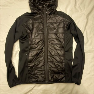Svart jacka från Cross Sportswear - Säljer en svart jacka från Cross Sportswear med dragkedja och huva. Jackan har en quiltad framsida och är tillverkad i syntetmaterial. Perfekt för kyligare dagar. Är i i princip nyskick och inga defekter.