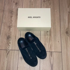 Svarta sneakers från Axel Arigato - Snygga svarta sneakers i mocka från Axel Arigato med dragkedja framtill. Perfekta för en stilren look. Kommer med originalkartong. Priset kan diskuteras så skicka ett förslag🌟