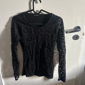 Svart spetsblus från Amisu - Elegant svart spetsblus från Amisu med långärmad design. Perfekt för en stilren look med vacker spetsdetalj som ger en feminin touch. Blusen har en figurnära passform och rund halsringning.