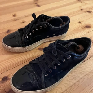 Mörblåa lanvin captoe - Snygga mörkblåa sneakers med snörning och vit sula. Skorna har en glansig finish och klassisk design som passar till många tillfällen.                  Ena skosnöret är kortare då tråden gått av och vänstra skon saknar og sula därför sänkt pris