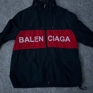 Svart och röd vindjacka från Balenciaga - Snygg svart vindjacka med röd panel och Balenciaga-logga i vitt. Jackan har dragkedja och huva, perfekt för en stilren look. Passar bra för både vardag och speciella tillfällen.
