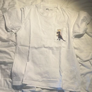 Jeans-michel BASQUIAT x Uniqlo - Säljer en vit t-shirt från Uniqlo med en cool dinosaurie och krona tryckt på bröstfickan. Designen är inspirerad av Jean-Michel Basquiat. Perfekt för dig som gillar konst och streetwear!