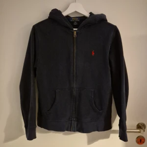 Mörkblå zip up från Ralph Lauren - Säljer en snygg mörkblå zip up från Ralph Lauren med dragkedja och huva. Den har en klassisk design med det ikoniska röda logotypen på bröstet och praktiska fickor framtill. Perfekt för en avslappnad stil.