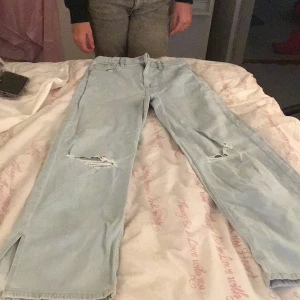 Ljusa jeans med slitningar - Säljer ett par ljusa jeans med slitningar på knäna och slitsar vid bensluten. De har en avslappnad passform och är perfekta för en casual look.