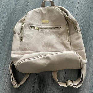 Beige/Rosa väska från Don Donna - Snygg beige ryggsäck från Don Donna. Väskan har flera fickor och justerbara axelremmar. Perfekt för vardagsbruk med sin rymliga design och snygga detaljer i guld 🥰