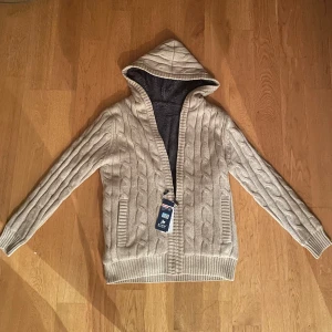 Beige stickad kofta med dragkedja - Mysig beige stickad kofta med dragkedja och huva från Maves. Tröjan har ett kabelstickat mönster och är fodrad med mjukt material för extra värme. Perfekt för kyliga dagar. Liten i storleken L men snarare som en M