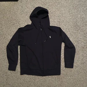 Mörkblå hoodie från Ralph Lauren - Säljer en stilren Mörkblå hoodie från Ralph Lauren med dragkedja och det klassiska logotypbroderiet på bröstet. Perfekt för en avslappnad look. Den har långa ärmar och en praktisk huva med snörning. Pris kan diskuteras