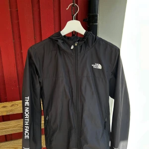 Svart vindjacka från The North Face - Säljer en svart vindjacka från The North Face med huva och dragkedja. Jackan har logotypen tryckt på bröstet och ärmen. Perfekt för blåsiga dagar. Ett litet hål vid vänster arm, men syns knappast.