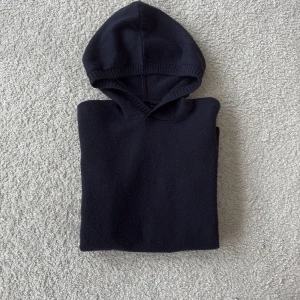 Marinblå stickad hoodie -  Tja! Säljer en stilren svart stickad hoodie med ribbade detaljer vid ärmslut och nederkant. Perfekt för kyliga dagar och ger en avslappnad look. Passar både till jeans och mjukisbyxor.  Nyskick Om du har frågor eller funderingar är det bara att fråga!