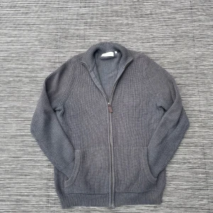 Grå stickad kofta med dragkedja - Säljer en stilren grå stickad kofta från Fredrik Anderson Copenhagen. Koftan har en praktisk dragkedja framtill och är tillverkad i ett mjukt material som passar perfekt för kyligare dagar. Den har långa ärmar och en ribbad struktur som ger en klassisk look. Tröjan är i storlek M medium🙌🙌 Om du har några frågor är det bara att höra av 😁👍