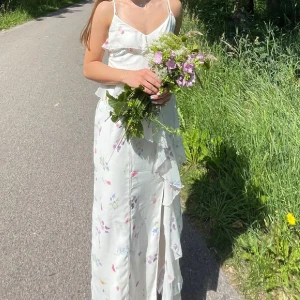 Vit blommig långklänning - Säljer en vacker vit långklänning med blommigt mönster. Klänningen har smala axelband och volangdetaljer. Perfekt för sommarens festligheter. Använd en gång, säljes då den är för liten 💕