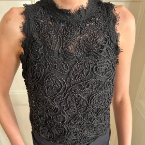 Svart spetsblus med dragkedja - Säljer en svart ärmlös spetsblus med hög hals och broderat mönster. Blusen har en synlig dragkedja i ryggen och är i spets. Är från Zara och i storlek S.