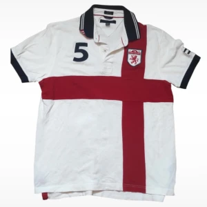 Tommy Hilfiger Chief Kief polo - Säljer en vit piké t-shirt från Tommy Hilfiger med rött korsmönster framtill, broderad sköld med lejon och siffran 5 på bröstet. Kragen är randig i svart och vitt med röda detaljer. Perfekt för en sportig look.