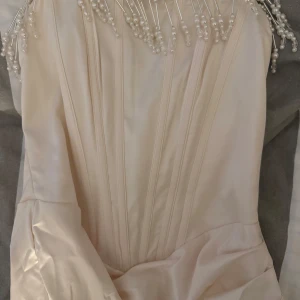 Champagnefärgad långklänning med pärldetaljer - Elegant långklänning i champagnefärgad satin med smala sömmar framtill och unik pärlprydd halsringning. Klänningen har en figurnära passform och är perfekt för bal eller festliga tillfällen.