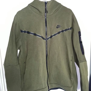 Nike tech fleece - Nike hoodie i olivgrönt med hel dragkedja och huva. Svart Nike-logga på bröstet och dekorativt band över bröstet. Ficka med dragkedja på ena ärmen och två sidofickor. Perfekt för en sportig och avslappnad stil.