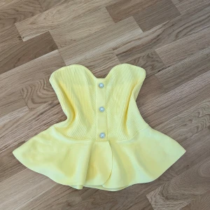 Gul korsettopp med pärlknappar - Säljer en söt gul korsettopp med volang nertill och dekorativa pärlknappar framtill. Toppen har en figurnära passform och är tillverkad i ett ribbat material som ger en snygg struktur.