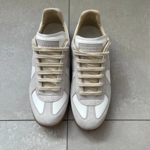 Maison Margiela sneakers i vitt skinn och mocka - Säljer ett par klassiska Maison Margiela sneakers med vit ovandel i skinn och ljusgrå mockadetaljer. Skorna har beige snörning och en brun gummisula. Perfekta för dig som gillar stilrena och tidlösa sneakers.