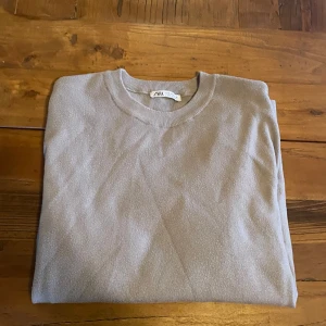 Beige t-shirt från ZARA - Säljer en stilren beige t-shirt från ZARA i storlek M. T-shirten har rund halsringning och är tillverkad i ett mjukt material som känns skönt mot huden. Perfekt för en avslappnad look. Plagget har blivit lite nopprigt.