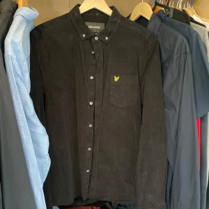 Svart manchester-skjorta från Lyle & Scott - Svart skjorta i manchester från Lyle & Scott med klassisk krage, knappstängning och bröstficka med gul broderad logga. Perfekt för dig som vill ha en stilren och tidlös look.