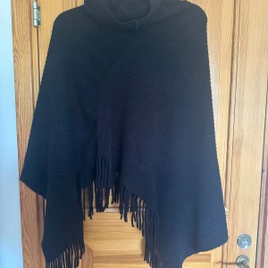 Svart poncho med fransar och polokrage - Svart poncho med polokrage och snygga fransar längs nederkanten. Perfekt att slänga över en tröja för extra värme och stil. Mjuk och skön, passar till många outfits.