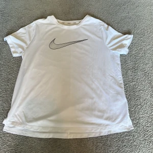 Vit t-shirt från Nike med svart logga - Vit t-shirt från Nike med klassisk svart swoosh-logga på bröstet. T-shirten har rund halsringning och korta ärmar. Perfekt för träning eller en sportig vardagslook.