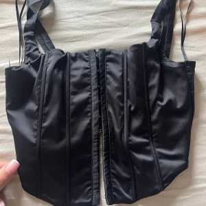 Svart korsettopp från Gina Tricot - Säljer en svart korsettopp från Gina Tricot med breda axelband och markerade sömmar framtill. Toppen har hakknäppning framtill och är tillverkad i ett glansigt material som ger en snygg siluett.