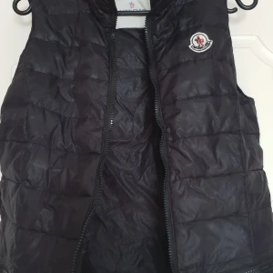 Svart dunväst från Moncler - Snygg svart dunväst från Moncler med klassisk logotyp på bröstet och dragkedja framtill. Västen har två fickor med dragkedja och quiltad design. Perfekt för lager-på-lager och stilren look.
