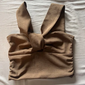 Brun croppad knyt-topp från H&M - Säljer en brun croppad topp från H&M med breda axelband och knytning framtill. Toppen är ärmlös och har en strukturerad yta i ett mjukt material. Perfekt för varma dagar eller som ett statement-plagg.