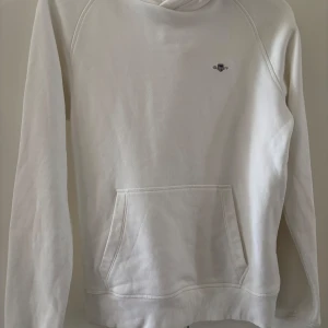 Vit hoodie från Gant - Säljer en stilren vit hoodie från Gant med broderad logga på bröstet. Tröjan har huva och en stor magficka framtill. Perfekt för dig som gillar klassisk och enkel stil.