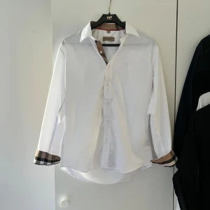 Vit skjorta med rutiga detaljer från Burberry - Säljer denna otroligt snygga burberry skjortan, den är i mycket bra skick typ 9,5/10, Inge defekter. Storlek L jag på bilden är 183 cm 68 kg, hör av er vid funderingar och pris kan diskuteras 