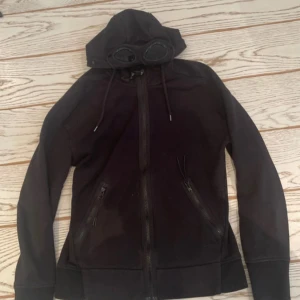 Svart hoodie med goggles från C.P. Company - Svart hoodie från C.P. Company med ikoniska goggles i huvan och dragkedja framtill. Jackan har två snedställda fickor med dragkedja och justerbar huva med snörning. Perfekt för dig som gillar streetwear och vill sticka ut.