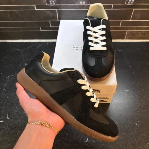 Svarta sneakers från Maison Margiela - Säljer ett par svarta sneakers från Maison Margiela med beige innersida och bruna gummisulor. Skorna har vita snören och är tillverkade i en mix av skinn och mocka. Klassisk design med rund tå och platt sula.