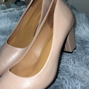 Beiga pumps med blockklack - Säljer ett par stilrena beiga pumps med rund tå och bred blockklack. Skorna har en klassisk design och är tillverkade i ett slätt material som ger en elegant look. Perfekta för dig som vill ha en enkel men snygg sko till många tillfällen.