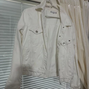 Vit jeansjacka från Tommy Hilfiger - Säljer en klassisk vit jeansjacka från Tommy Hilfiger med silverfärgade knappar och bröstfickor. Jackan har lång ärm och diskret logga på ena fickan. Perfekt för att lyfta din outfit med en fräsch och stilren look.